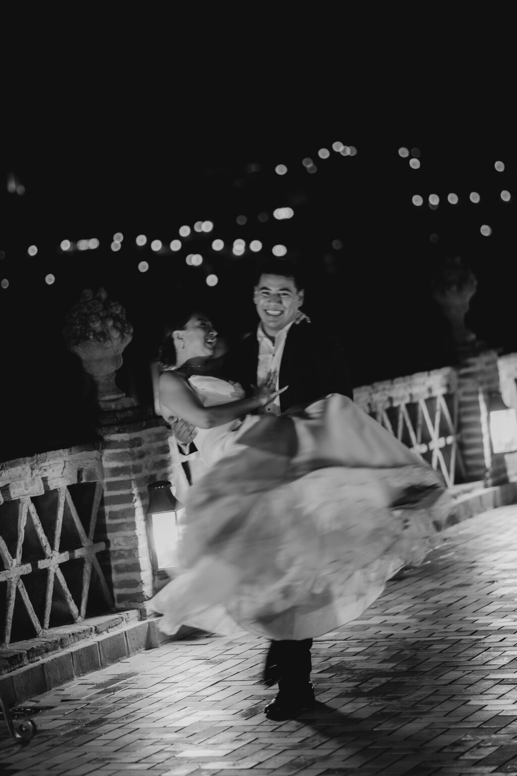 Fotografía en blanco y negro de un novio llevando en brazos a la novia durante la celebración de su boda de noche en Alicante, realizada por Castizzo, fotógrafo de bodas.
